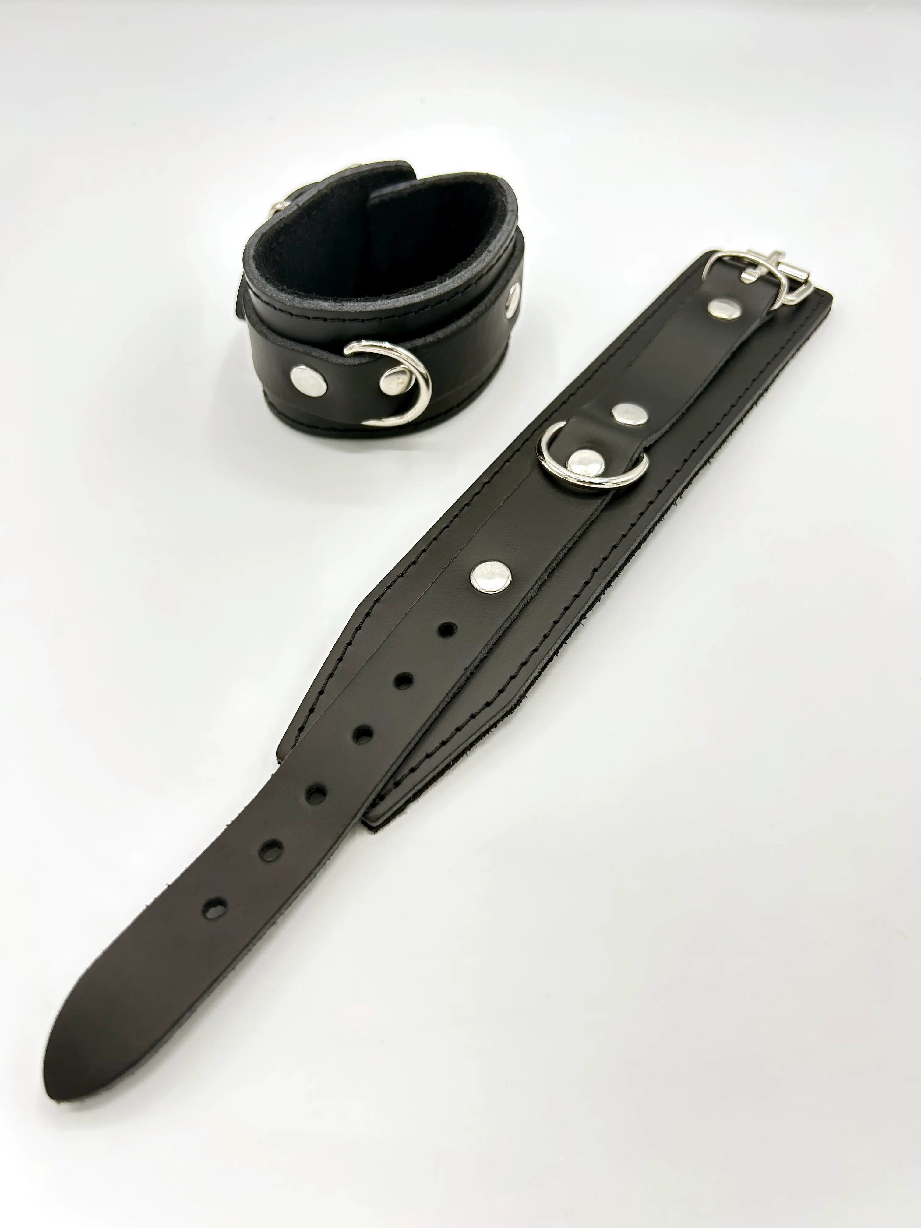 Zwei schwarze Leder-Handfesseln mit D-Ring, Nieten und schwarzem Innenfutter, eine geschlossen und eine aufgeklappt auf weißem Hintergrund